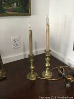Two brass candlesticks electrified as lamps sitting on dark wooden surface against white wall.
