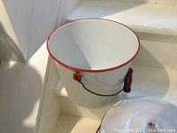 White enamelled pot with red rim and wooden handle inside, sitting on a white step.