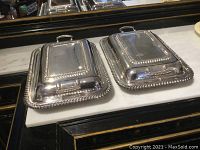 Two silver on copper rectangular entree dishes with lids, dragooned edges, and handles shown on marble surface.
