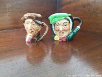 Two Royal Doulton character mugs, one of an elderly lady with green headscarf and one of a working class Englishman with a flat cap, on a wooden surface.