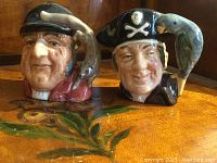 Two Royal Doulton character jugs side by side on floral surface, depicting a Huntsman and a Pirate with detailed painted designs.