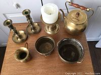 Full top view of the six brass items on wood surface including candlesticks, teapot with wooden handle, jug, footed pots, and candle stand.