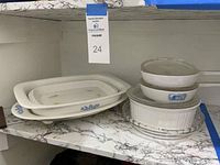 Photo showing multiple Corning Ware dishes stacked on a shelf including rectangular serving trays, round casserole dishes with glass lids, and a pie plate.