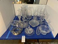 Overview of many of the glass items arranged on a blue cloth including candle sticks, vases, glasses, bowls, and serving dishes