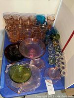 Various pink depression glass glasses, bowls, clear serving platters and colored glass pieces arranged on a blue surface.
