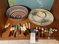 Wide angled photo showing the assortment of 17 cheese spreaders with various holiday and novelty handles arranged in front of the chip and dip platter and the striped Havana bowl on a wooden shelf.