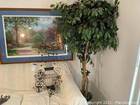 View showing framed landscape art, two metal candle sconces attached below, and a tall artificial tree in woven basket next to a white couch