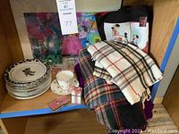 Shelf with stack of eight white ceramic plates with black rim design, Royal Grafton cup and saucer with floral pattern, and four folded plaid scarves in different colors plus small decorative items
