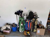 Photo showing several golf bags including green, blue Florida Gators, and beige with assorted golf clubs and boxes of golf balls and sports equipment on floor.