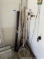 Photo showing multiple fishing rods with reels leaning against a wall along with fishing nets on the floor.