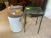White metal barrel table with natural wood round top and rope handles next to green metal square table with distressed finish on tile floor