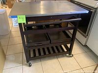 Front and side view of kitchen island cart showing black painted frame, two drawers, towel bar, trays on middle shelf, and casters.
