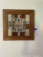 Full view of the square Elgin wall clock featuring a wood frame and stained glass sections in earth tones with the clock face centered showing Arabic numerals.