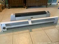 Two portable space heaters placed on floor side by side, one black-gray (Lasko) and one white (Slant/Fin)