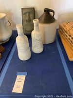 Photo showing two white glass bottles with painted black designs, crock jug with dark brown top and handle in background, and mask wall decor behind them.
