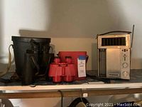 Shelf view showing black Sard binoculars on left in leather case, red child's binoculars centered with carrying case behind, and vintage Windsor radio on right.