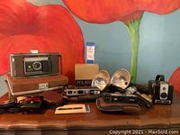 Overview of vintage cameras and accessories including Polaroid camera in case, Kodak Instamatic, Minolta 450 E, Brownie Hawkeye, flash attachments, and flash bulbs.