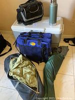 Photo showing large white Coleman cooler, Stanley thermos, and tents in carrying bags with blue folding seats stacked on cooler