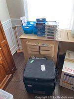 Wide view showing a black fabric rolling suitcase Crop in Style on the floor next to a beige desk and drawers, with plastic containers holding sewing supplies on top of the desk.