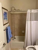 Photo of bathtub with installed white and translucent shower curtain, blue towel on rod, beige tiled wall, and framed blue sky beach scene art.