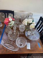 Table display of clear pressed and cut glass items including canisters, vases, jars, plates, and decor with floral and geometric motifs.