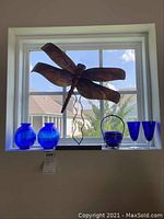View of metal dragonfly wall art mounted above windowsill, with six cobalt blue glass items displayed on sill under natural light.