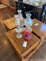 Three antique pressed glass oil lamps arranged on wooden tables with a kitchen in the background. One oil lamp has red glass base and etched chimney with scenes. The others have clear chimney glass with distinct shapes and detailed bases.