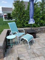 Full view of turquoise mesh patio chair with matching ottoman and small blue metal table on stone patio with greenery background