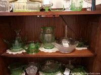 Front view of the first shelf showing multiple green uranium and clear depression glass items, including bowls, a vase, and a citrus juicer.