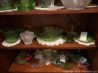 Second shelf of a wooden display cabinet holding various green uranium glass and clear glass depression glass pieces including a pitcher, sugar bowl, relish bowls, and plates.