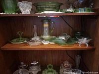 Wide view of a wooden shelf holding assorted uranium glassware including plates, bowls, teapot, and small dishes.