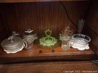 Shelf of assorted glassware including uranium glass handled serving tray and other covered bowls, pitchers, and dessert plates.