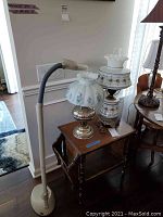 Two hurricane lamps with floral glass shades on wooden table beside an adjustable work light.