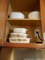 Upper cabinet with stacked vintage CorningWare casserole dishes with orange, yellow, and fruit flower pattern and white mixing bowls.