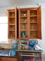 Open kitchen cabinet with multiple glassware items including glasses and pitchers on countertop and inside cabinets.