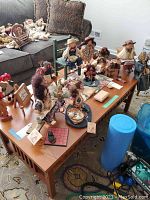 Photo showing multiple Lizzie High dolls arranged on and around a wooden coffee table, with several wooden furniture pieces including chairs and benches visible, along with small wooden carved animals and homespun figures on the coffee table.