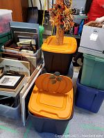 Photo showing three large plastic tubs with orange lids stacked and placed among frames, containing Halloween and fall-themed decor.