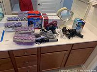 Photo shows two sets of CONAIR electric hair rollers, assorted hairdryers, WAHL clip and trim kit in box, illuminated mirror, and Vicks steam inhaler box on a bathroom counter.