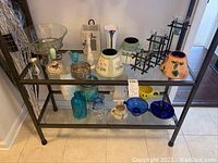Wide view of glass and decorative items on glass-top table showing vases, bowls, candleholders, lampshades and decorative bottles.