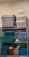 Grey large plastic storage containers and white translucent drawer units on top shelf, medium blue and teal containers and small clear containers on middle shelf, various smaller blue, green, and red containers stacked on bottom shelf.