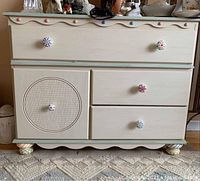 Front view of wood dresser showing top large drawer, lower right two smaller drawers, and left cabinet door with round textured detail. Painted cream with pastel trim and colorful ceramic knobs.