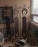 Three-panel wrought iron and woven room divider leaning against a wall with framed artwork in the background.