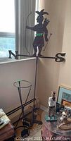 Wrought iron weather vane shown resting on a window ledge; archer silhouette with bow and arrows and directional indicators N and S visible.