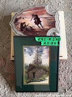 Two framed art pieces on top of a stack of books: carved white frame with mounted warrior scene and black-framed print beneath