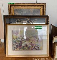 Three framed artworks standing one behind the other, main watercolour floral painting in a gold frame in front, a poster with gold script text behind it, and a decorative ornate gold frame artwork in the back.