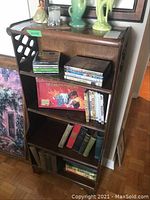 Front view of wooden bookcase showing three shelves filled with books and media, decorative side panels visible, top shows ring marks and wear.