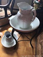 Full view of two white ironstone washbasin and pitcher sets arranged on a black iron folk art stand on wooden floor with furniture in background.