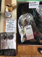 Gold-tone designer cuff bracelet signed Alisha dated 1990, placed in box with tissue and packaging visible. Adjacent black tray holding assorted beaded necklaces and other jewelry under plastic bag labeled beaded necklaces.