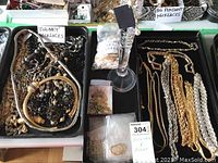 Photo showing two trays with an assortment of chunky necklaces, chains, faux pearls, and bags of small jewelry parts and a jewelry display stand in the center.