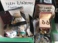 Photo showing two boxes filled with assorted earrings, one labeled new costume earrings and the other labeled post earrings with backs, containing various dangling and hoop styles predominantly gold-tone and silver-tone with some bead accents